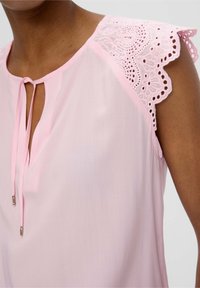 Femme portant un chemisier sans manches rose pâle avec des détails en dentelle sur les épaules et une petite ouverture en forme de goutte fermée par de fines cordelettes.