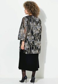 Schwarze und metallische florale Überjacke mit Dreiviertelärmeln, aufwendiger Stickerei, kombiniert mit einem schwarzen Rock und hohen Schuhen.