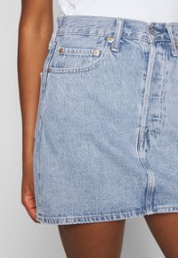 Jupe en denim bleu clair à taille haute, avec poches avant et couture latérale. Le tissu est légèrement texturé, avec un léger effet délavé et des détails de couture.