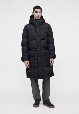 Jeune homme portant un long manteau matelassé noir avec une capuche, pantalon gris et baskets noires, debout devant un fond clair uni.