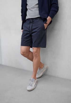 Hombre con pantalones cortos azul marino, camiseta gris, chaqueta azul marino y zapatillas gris claro, de pie con las manos en los bolsillos contra una pared clara.