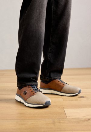 Asmuo, vilkintis juodas kelnes ir šviesiai rudas batų su raišteliais, stovi ant medinės grindų.