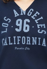 Chemise bleu marine à col en V avec un texte bleu clair indiquant "LOS ANGELES 96 CALIFORNIA" et en dessous, en cursive plus petite, "Paradise City".