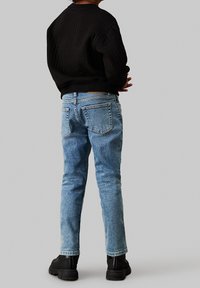 Persona con un suéter negro, jeans azul claro y botas negras de pie contra un fondo gris liso, de espaldas.