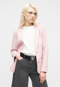 Γυναίκα με ξανθά μαλλιά, που φοράει ένα ανοιχτό ροζ σακάκι, λευκό πουκάμισο, σκούρα γκρι τζιν και μια μαύρη ζώνη με ασημένια αγκράφα σε σχήμα καρδιάς.