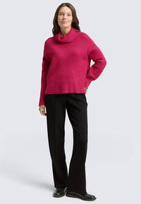 Pull rose oversize à côtes avec un large col bénitier, associé à un pantalon noir à jambes droites et des chaussures noires, sur un fond clair.