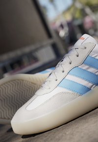 Hvid sneakers med lyseblå striber og beige ruskind detaljer, vist tæt på en betonoverflade med sløret baggrund.