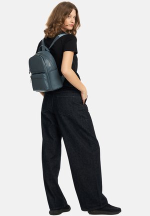 Lichtblauwe leren rugzak met voorvak met rits, gevormde contour, verstelbare banden, gedragen door een persoon in een zwart T-shirt en donkere broek.