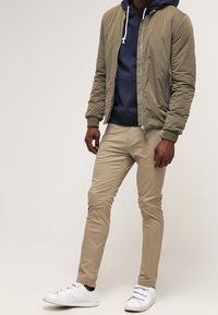 Man med olivgrön bomberjacka, marinblå hoodie, beige slim-fit byxor och vita sneakers, som står mot en enfärgad ljus bakgrund.