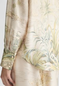Blouse à manches longues en tissu satiné avec un fond beige et des imprimés botaniques verts et des accents clairs, poignets détaillés avec des boutons.