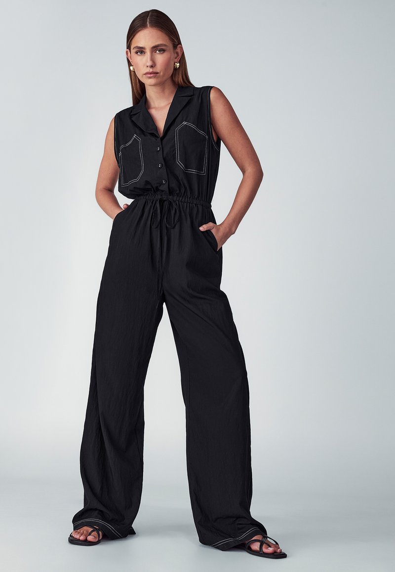 Ženska stoji z rokama v žepih, oblečena v brezrokavni črn kombinezon z vrvico v pasu, širokimi nogami, sprednjimi žepi in črnimi sandalami.