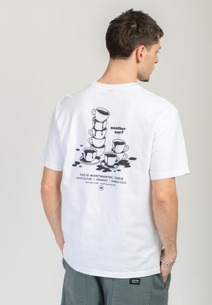 Mężczyzna w białej koszulce z czarnym nadrukiem przedstawiającym ułożone na sobie filiżanki do kawy i rozlaną kawę, z napisem „kolejny?” oraz odniesieniami do paryskiej kawiarni.