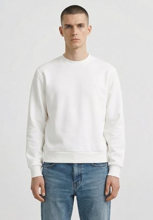 Giovane uomo con capelli corti che indossa una felpa bianca semplice e jeans blu, in piedi di fronte a uno sfondo grigio neutro.