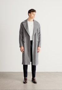 Γκρι παλτό από μαλλί με διπλό στήθος, μεγάλες πέτο και πλαϊνές τσέπες. Φορεμένο πάνω από άσπρο πουκάμισο και μαύρο παντελόνι, ολοκληρωμένο με καφέ παπούτσια.