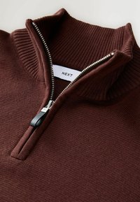 Hnedá mikina na zips s vysokým golierom, s rebrovanou textúrou a čiernym ťahákom na zips. Na štítku je napísané "NEXT." Tkanina vyzerá mäkko a textúrovane.