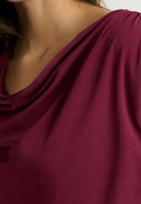 Cowl-neck top i burgunderfarvet stof, der har en blød, draperet tekstur med milde folder. Nærbillede fremhæver det glatte materiale.