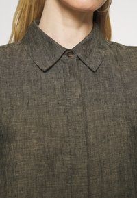 Tummanvihreä pellavapaitapusero, jossa on napillinen kaulus ja hienovarainen rakenne. Sisältää keskipaneelin, jossa on nappiyksityiskohta ja kontrastivärjäys.