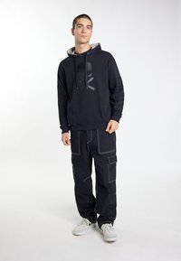 Zwarte hoodie met logo, voorzien van een trekkoord en grijze voering, gecombineerd met oversized zwarte cargo broek met witte stikdetails.