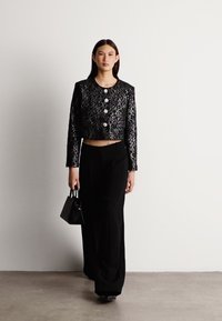 Giacca nera accorciata con un motivo floreale testurizzato e bottoni rotondi in argento, abbinata a pantaloni neri a vita alta e una piccola borsa.