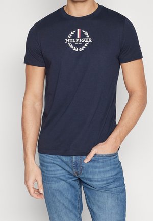 Navy blue cotton t-shirt with short sleeves, featuring a white circular logo and laurel wreath design with red and white stripes.