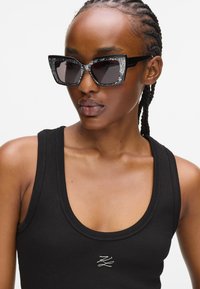 Μαύρα γυαλιά ηλίου cat-eye με γυαλιστερές λεπτομέρειες στη στεφάνη και τόνωση στους φακούς. Το μοντέλο φοράει ένα μαύρο ριγέ tank top με λεπτομέρεια λογότυπου.