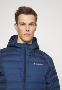 Giacca isolante blu navy con zip frontale e cappuccio. Caratterizzata da baffi orizzontali e un piccolo logo bianco Columbia sul petto.