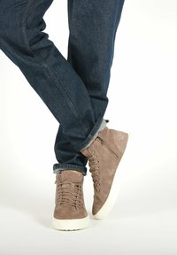 Brune ruskind høj-top sneakers med en hvid gummisåle, der har en lynlås i siden, snørebånd foran, samt denim jeans med opkrøllede muffer.