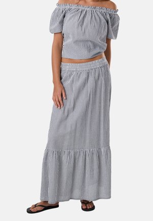 Frau trägt ein zweiteiliges Outfit mit grauen und weißen vertikalen Streifen, bestehend aus einem schulterfreien Crop-Top und einem langen, gestuften Rock, kombiniert mit schwarzen Flip-Flops.