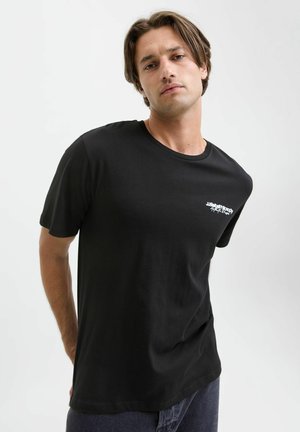 Joven con cabello de longitud media que lleva una camiseta negra de cuello redondo con texto blanco en el pecho, sobre un fondo claro y sencillo.