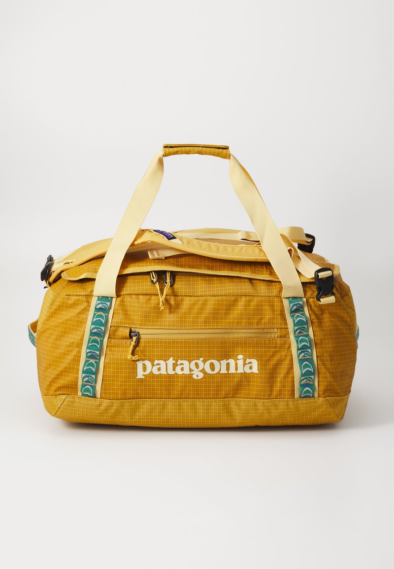 Žltá taška Patagonia s textúrovanou tkaninou, prednou zipsovou kapsou a zelenými vzorovanými popruhmi. Obsahuje pevné rukoväte a potlač loga.
