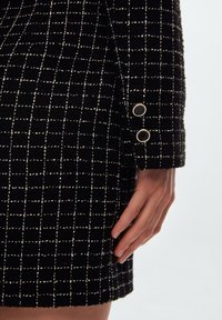 Zwart gestructureerde stof met een rasterpatroon, met een manchetdetail met drie knopen in zwart en goud. Close-up van mouw en hand.