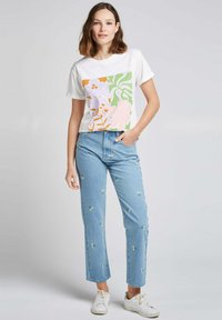 T-shirt blanc avec un grand motif floral en rose, vert et orange, associé à un jean bleu clair avec broderie florale et des baskets blanches.
