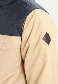 Chaqueta en tonos marrón y gris con tejido texturizado, que cuenta con un bolsillo en el pecho, cierre de botones y un parche del logo de Vans en cuero marrón en el hombro.
