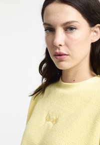 Gele fleece sweatshirt met geribbelde halslijn, voorzien van een reliëf-vleermuislogo op de borst en een gestructureerd oppervlak. Model afgebeeld van de schouders omhoog.