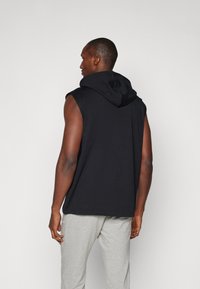 Schwarzer ärmelloser Hoodie mit großer Kapuze, aus weichem Material und lockerem Schnitt. Kombiniert mit hellgrauen Jogginghosen. Schlichte Gestaltung, ohne Muster.
