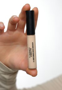 Beige concealer tube med svart hette, med trykt tekst. Tuben har en sylinderform og holdes mellom fingrene mot en lys bakgrunn.