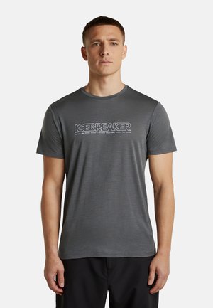 Mann trägt ein eng anliegendes dunkelgraues Kurzarm-T-Shirt mit "ICEBREAKER" Logo in weißem Text auf der Brust, steht vor einem schlichten Hintergrund.