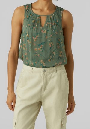 Blouse verte fleurie sans manches avec un décolleté en trou de serrure, portée avec un pantalon beige clair taille haute avec poches.
