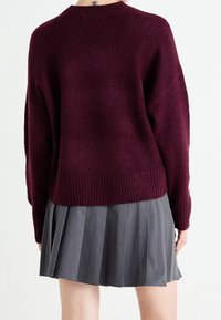 Pull en maille bordeaux avec ourlet côtelé, manches larges et encolure ronde, associé à une jupe plissée grise. Tissu texturé visible.