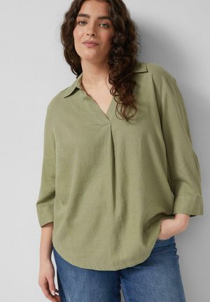 Vrouw met lang krullend haar, gekleed in een losse olijfgroene blouse en blauwe spijkerbroek, staand met één hand in de zak tegen een grijze achtergrond.