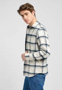 Camisa de franela beige con un patrón de cuadro en azul marino y blanco, con botones en el frente, cuello tipo gala y mangas largas; combinada con jeans azules.