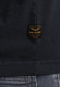 Camiseta negra de algodón con una etiqueta rectangular con texto dorado y alas, situada cerca del dobladillo. Se ve tela de mezclilla gris por debajo.