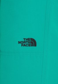 Turkoosin värinen kangas, jossa on sileä tekstuuri ja mustalla kirjailtu "The North Face" -logo. Suunnittelussa on tasku- ja ompelutaitteita.