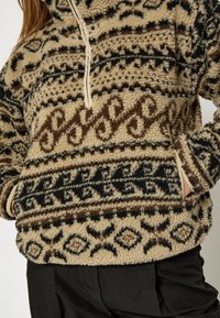 Béžový fleecový pulóver s tmavohnedými a čiernymi geometrickými vzormi, s polovičným zipsom na krku a prednou vreckom. Mäkká, textúrovaná tkanina.