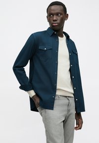 Jonge man in een knopen navy shirt over een beige sweater en grijze jeans, staand met een hand in zijn zak tegen een witte achtergrond.