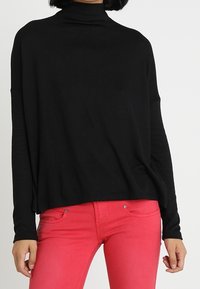 Pull à col roulé noir à manches longues, coupe décontractée, associé à un jean rose avec poches et détails de boutons. Texture en maille douce.