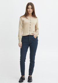 Beige gebreide cardigan met knoopsluiting, gedragen over donkerblauwe skinny jeans. Zwarte leren instappers maken de outfit af. Eenvoudig ontwerp, casual stijl.