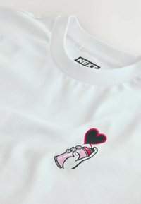 Weißes T-Shirt mit einem kleinen, bestickten Motiv einer Hand, die eine rosa Sprühdose hält, aus der ein schwarzes Herz mit rotem Rand freigesetzt wird.