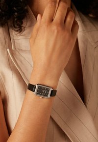 Rechthoekig zilveren polshorloge met een zwarte wijzerplaat, witte uurmarkeringen en een zwarte leren band, gedragen aan een lichtgekleurde hand tegen een beige achtergrond.