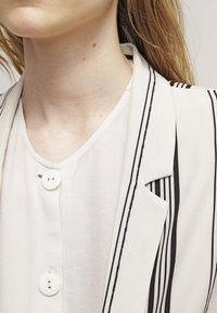 Bílý blazer s černými pruhy, zdobený klopami a uzávěrem na jeden knoflík, vrstvený přes lehkou, texturovanou blůzu s detaily knoflíků.